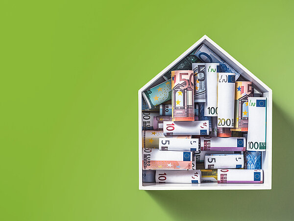 in einem hausförmigen Bilderrahmen, der an einer grünen Wand hängt, sind aufgerollte Geldscheine gestapelt und sortiert